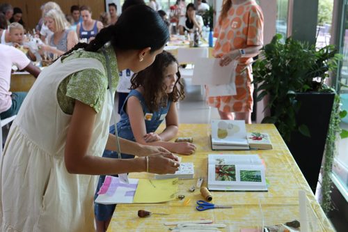A younger woman talking to a girl, they are both standing at a table which is laid with paper and books illustrated with pictures of plants
