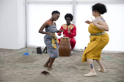 Two female performance artists in Ghanian costume dance on the sand-covered floor of Senga Nengudi's 'Sandmining' installation. Accompanying them with traditional Ewe drumming is Nii Kwartey Owoo, Director of Miishe African Music and Dance. He is sat by a large upright drum, which he is playing with one open hand and a stick in the other.