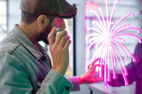 A man holds an audio device to his ear while touching a pink-lit acrylic panel illuminated with a dahlia firework with arching trails.