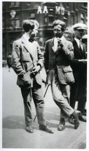 A vintage black and white photo showing two young men in their early 20s. They are dressed in dapper 1920s suits, one is leaning on a cane and smoking a pipe, the other is sporty a natty bow tie.