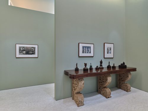 Eight small bronze sculptures by artist Henry Moore, displayed on a wooden table. Three framed drawings, also by Moore, are on the walls behind.