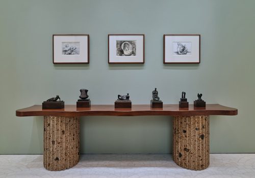 Six small bronze sculptures by artist Henry Moore, displayed on a wooden table. Three framed drawings, also by Moore, are on the wall behind.