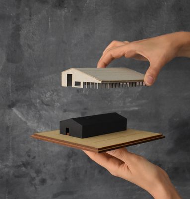 One hand holds a black model depicting the current gallery, on a wooden base; above it, a second hand holds a white model showing the redevelopment, hovering over the black model.
