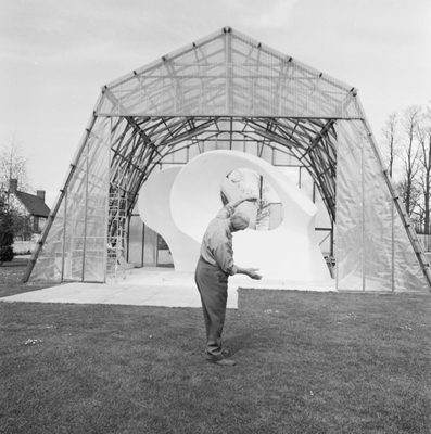 A black and white archive photo showing an older man standing in front of a large white sculpture. The sculpture takes the form of two ovals and the man is mirroring the shapes with his arms.