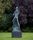 A colour photo showing a tall, abstract sculpture in an organic shape. It is standing on a plinth in a garden.