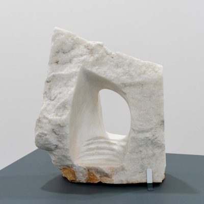 A white stone sculpture with a window-like opening carved through the middle, with shallow steps leading into it. The exterior is rough, while the interior is completely smooth.