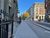 A pedestrian pavement with black railings and an old building with pillars on the left and a cycle lane on the right. A road junction is ahead, with an autumnal tree on the other side.