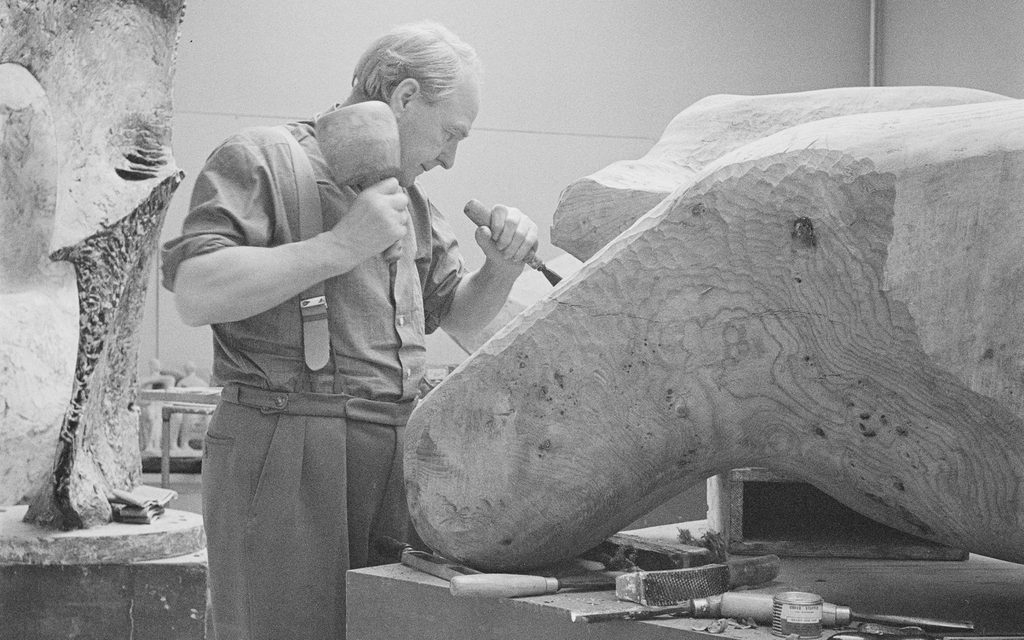 A black and white photo showing an older man in shirt and tie working with a mallet and chisel to carve a sculpture from a large piece of wood.