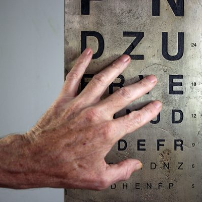 A splayed hand touching a bronze plate with lines of single black letters recessed into it. The letters are from a Snellen eye test chart and reduce in size from top to bottom.