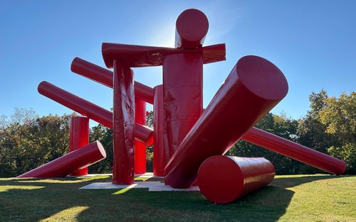 A large piece of outdoor sculpture, made from metal cylinders painted red and piled together in a semi-haphazard way.