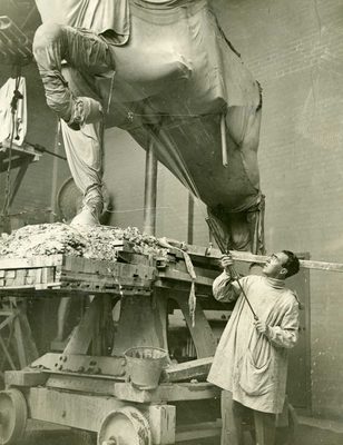 Vintage photo of a man spraying water upwards at a monumental sculpture of a horse, which is covered in sheets and resting on top of a large wooden platform with wheels.