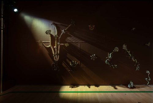 A brown gallery wall, on which several abstract metal sculptures are displayed; some look like arms, others are harder to make out. They are murkily lit from a single spotlight in the top left of the image.