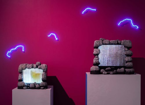 Two TV screens on plinths, showing static screens, framed with fake rocks. Behind, the wall is bright pink, with four squiggly blue neon tubes arranged around the TVs, mimicking electricity sparks.