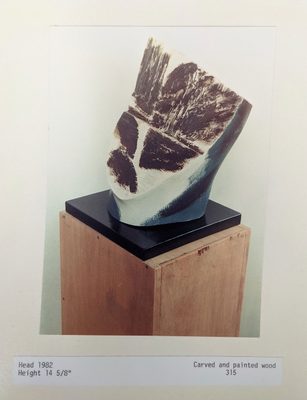 Abstract sculpture of a head by Anthony Hatwell, carved from wood and painted white, black and dark blue. The 'face' is very flat, with rough blocks of black paint giving the impression of hair, eyes and a mouth.