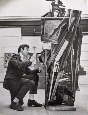 Black and white photo of a man wearing a suit, holding a small hammer, at work on a sculpture made from pieces of wood nailed together haphazardly.