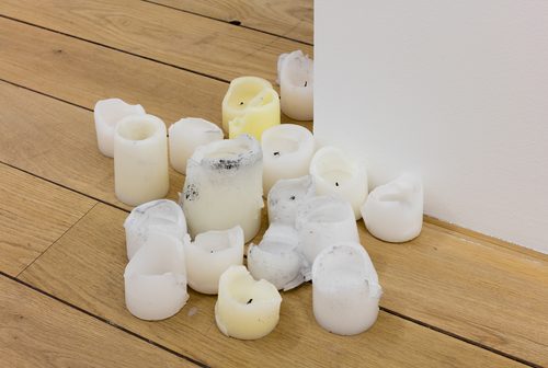 A cluster of sculptures of candles in various stages of being burnt sit on a wooden floor around the corner of a white wall