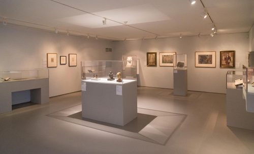 Wide shot of a gallery space, with framed drawings on the walls and small sculptures and books in display cases.