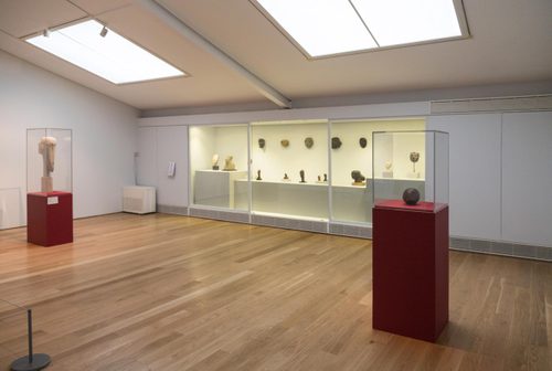 Wide shot of a gallery space, with two sculptures of heads on covered plinths, and a display case set into the wall holding small sculptures of heads and masks.
