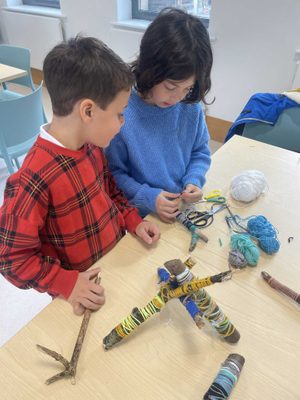 Two children at at table, wrapping sticks with wool to make small sculptures.