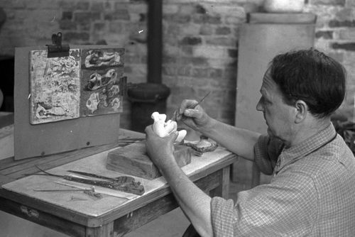 A black and white photo showing sculpture Henry Moore working on a plaster maquette in his studio in 1947. Moore is wearing a check shirt and is seated at a wooden work bench. On the bench, an open sketchbook is visible.