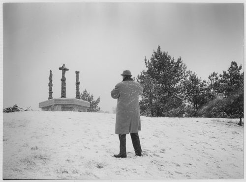 Black and white photo of a man wearing a smart coat and hat, seen from behind, taking a photo of abstract sculptures in a snowy field.