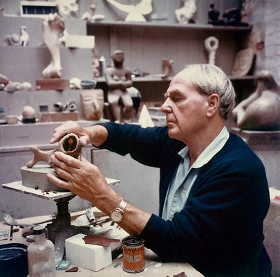 A vintage colour photo showing sculptor Henry Moore working in his maquette studio. He is moulding a small sculpture in some kind of clay.