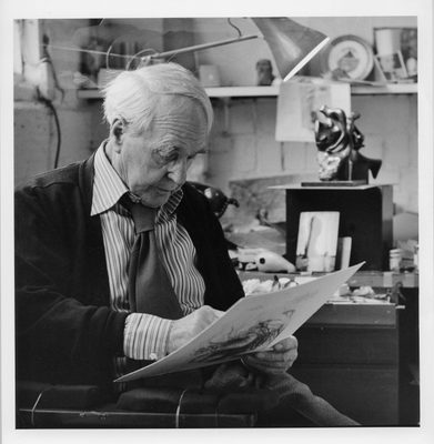 A black and white photo showing an elderly Henry Moore working on a drawing in one of his studios in 1982. Moore is wearing a jacket, striped shirt and tie with a fat knot. Behind him a small bronze sculpture of a mother and child is visible.