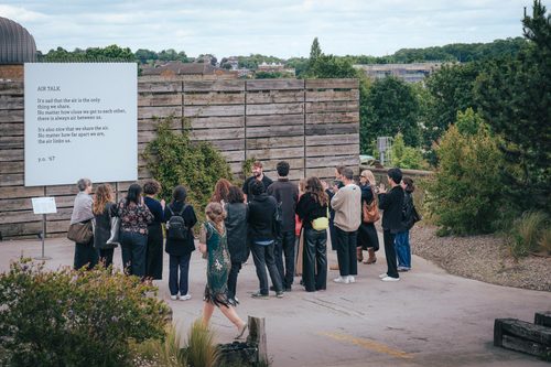 A group of people stand around a tour guide looking at a square board with a poem on it. The poem reads: 