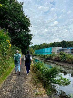 Two people walking along a canal path, with trees to their left, and large metal storage sheds on the other side of the canal to the right.