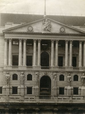 Black and white photo of the Bank of England, a large, seven storey building. The front has been styled to resemble classical Greek or Roman buildings, with ornate columns holding up the roof. Several figurative sculptures adorn the lower part of the building, situated between the windows, each standing around eight feet tall.
