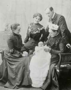 Black and white photo of a baby in a long white dress, surrounded by three women in dark, high-necked long dresses and a man wearing a suit.