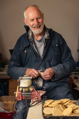 A man holding a plaster digestive biscuit and a carving tool in his hands. In front of him is a large pile of finished plaster biscuits and a large desk vice.