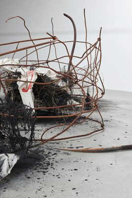 Detail of an abstract sculpture, with burned materials and rusty chicken wire at the centre, with rusted metal bars bent around it. The tip of a rusty harpoon lies on the floor just in front of it.
