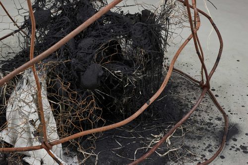 Detail of an abstract sculpture, with burned materials and rusty chicken wire at the centre, with rusted metal bars bent around it.
