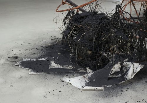 Close up view of tangled, matted fibres and fabric, stained black, attached to a rusted metal frame. It is covered in charcoal powder, making it look like it has been burned.