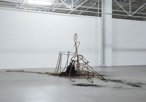 Abstract sculpture in a white gallery space. It is made from rusted metal harpoons and rope, arranged in a vaguely conical shape.