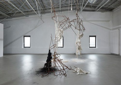 Two abstract sculpture in a white gallery space. One is conical in shape and sits on the floor, made of rusted metal hooks and fabric that looks to have been burned. The other is suspended from the ceiling, a mesh of twisted, rusted metal with matted white fabric hanging off it.