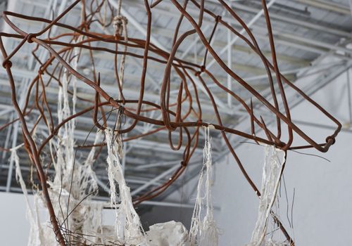 Close up view of part of a sculpture, showing the tips of several rusted iron hooks linked together to make a mesh, with strands of white fabric hanging off it.