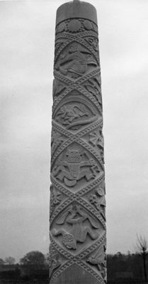 Black and white photograph of Eric Kennington's sculpture 'Comet Racer' (1936). The photo shows a pillar, made for the car park of the Comet Inn, Hatfield. The pub was built in brick in the shape of an aeroplane and faced an aircraft factory. The pillar depicts various means of flight from mythology, such as Icarus, a witch on a broomstick, and flying fish.