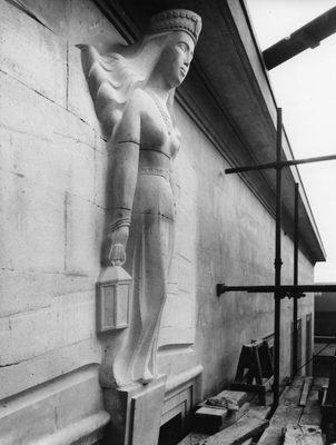 Black and white photo of a sculpture of a woman holding a lantern. She is jutting out from a sandstone wall, where she has been carved from the same blocks that make up the building.