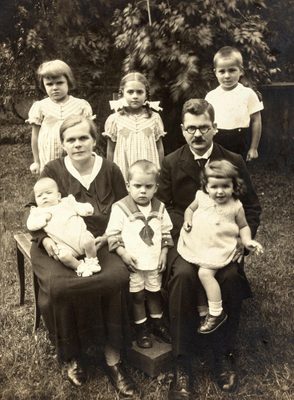 Black and white family portrait. The parents are seated, each with a child on their lap, and a small boy between them. Behind stand a row of three young children, two girls and a boy.