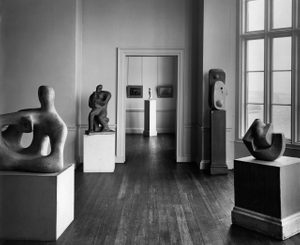A black and white photo of a sculpture exhibition displayed in a gallery. In the first room, nearest the camera, four large carvings are displayed on plinths. Visible through a doorway, in the next room, are two pictures hanging on the wall and one further sculpture.