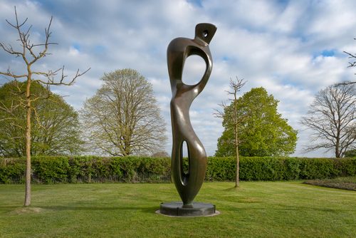 A tall, bronze sculpture of an abstract standing figure by Henry Moore, displayed on a manicured lawn in an English sculpture garden.
