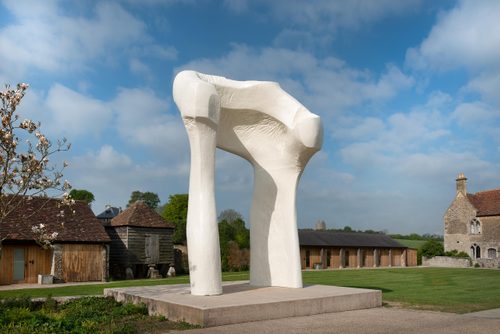 A large, arch-shaped sculpture by Henry Moore, displayed on the grounds outside some traditional English country buildings. The arch's shape and chalk-white colour gives the impression of bones.