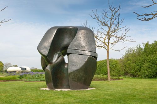 A large, bronze sculpture by Henry Moore, made of two interlocking pieces reminiscent of the joint between two bones. It is displayed on a manicured lawn in an English sculpture garden.