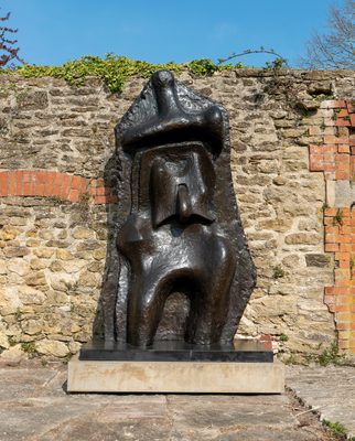A large, bronze sculpture of an abstract figure by Henry Moore, standing in front of an old stone wall that has been repaired with bricks.