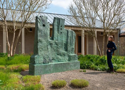 A woman looking at a large bronze sculpture by Henry Moore. The sculpture is abstract, a series of squat rectangular shapes punctuated with circular holes and horizontal and vertical lines, and has oxidised to a green colour.