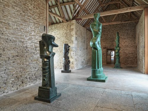 Four tall, bronze sculptures by Henry Moore, appearing to be somewhere between figurative sculpture and pillars. They are displayed in an old stone building that has been converted into a gallery.