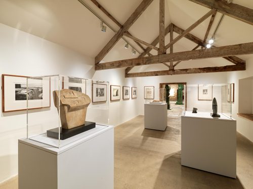 A white-walled gallery space with exposed wooden beams, showcasing work by Henry Moore. Framed drawings are hung on the wall, and there are small sculptures displayed underneath perspex containers on three plinths.