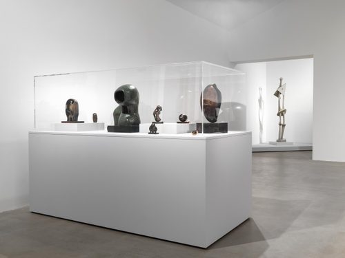 A white walled gallery space, with a large plinth displaying several small bronze sculptures by Henry Moore. Behind the plinth, there is a tall, spindly sculpture of a abstract standing figure.